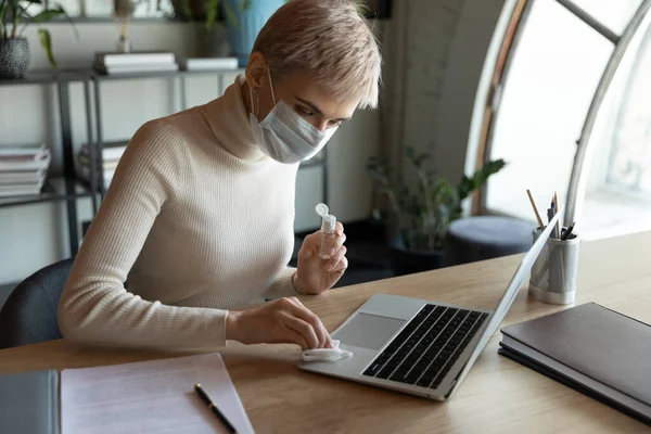 Sorumlu genç kadın tıbbi koruyucu yüz maskesi takıyor. Bilgisayarı dezenfekte ediyor.. — Stok fotoğraf