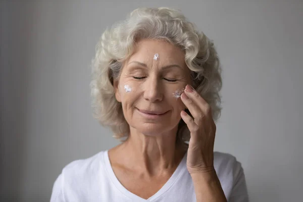Smiling beautiful middle aged senior retired woman nourishing moisturizing face. — Stock Photo, Image