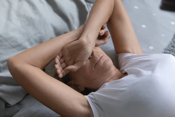 Senior woman covering eyes with arms, trying sleep in bedroom.