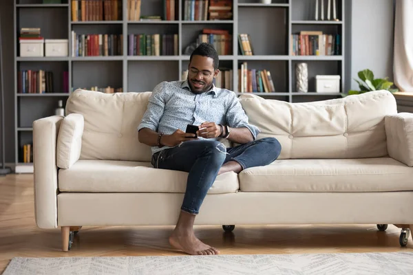 Smiling young african man sitting on comfy couch, playing game.
