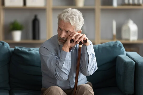 Tired upset mature man holding walking stick, sitting on couch — Stock Photo, Image