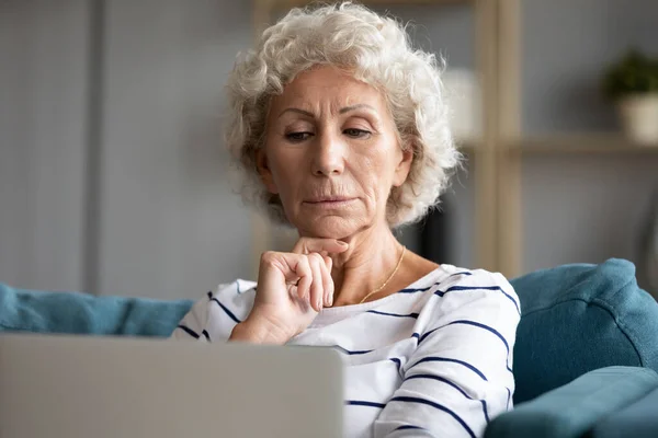 Serious pensive older woman looking at laptop screen