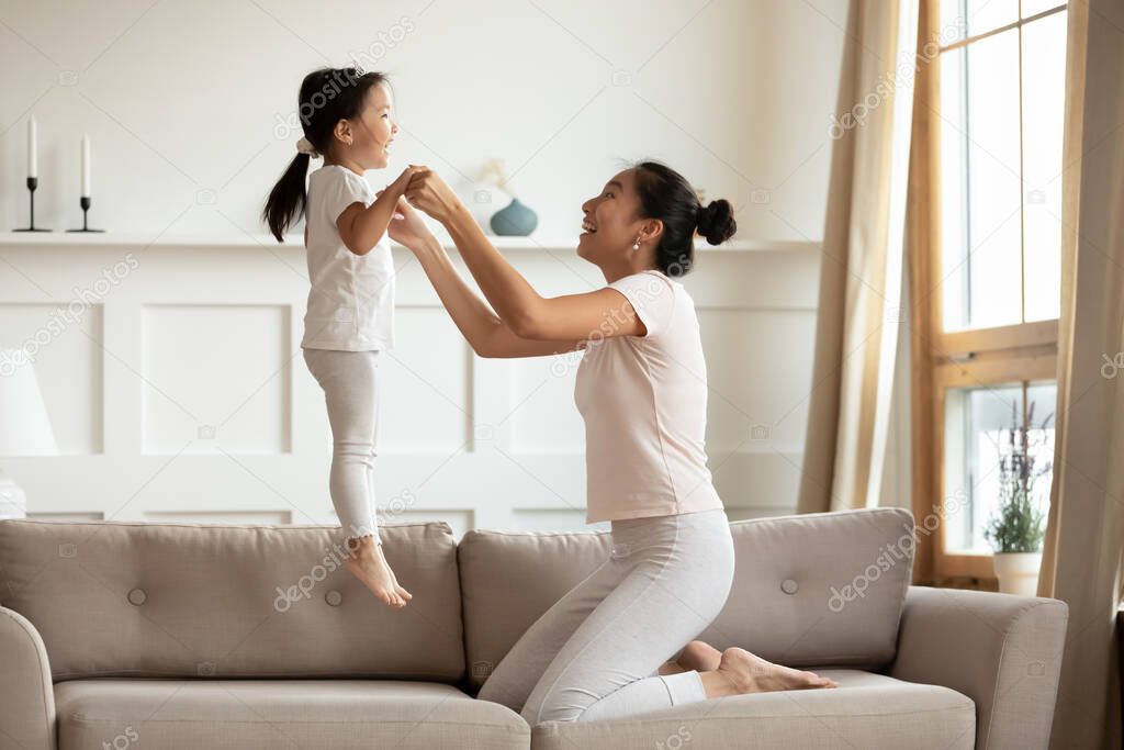 Mother holding hands little daughter while she jumping on couch