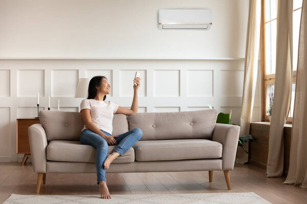 Asian woman switching on air conditioner while resting seated indoors