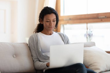 Afrikalı Amerikalı kadın dizüstü bilgisayarda çalışıyor, kanepeye oturuyor.