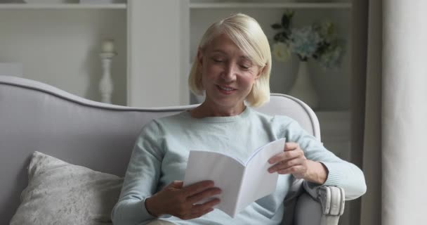 Glad mogen äldre kvinna vilar på soffan, läsa bok. — Stockvideo