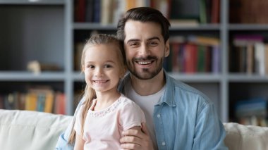 Baba ve kızı kanepede oturmuş kameraya bakarak gülümsüyor.