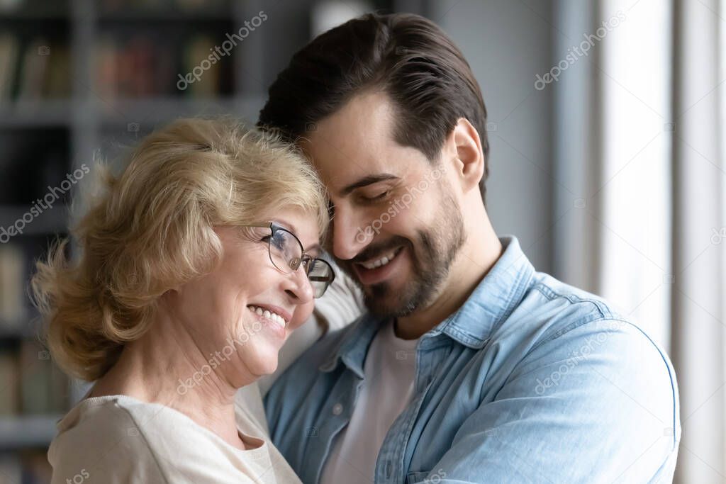 Millennial son and elderly mother hugging standing indoors