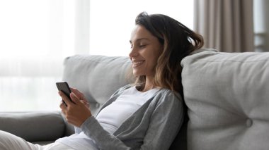 Telefonla gülümseyen kadın, evdeki rahat koltukta dinleniyor.