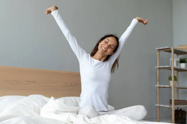 Wanita muda yang bahagia merentangkan tangan setelah bangun, duduk di tempat tidur — Stok Foto