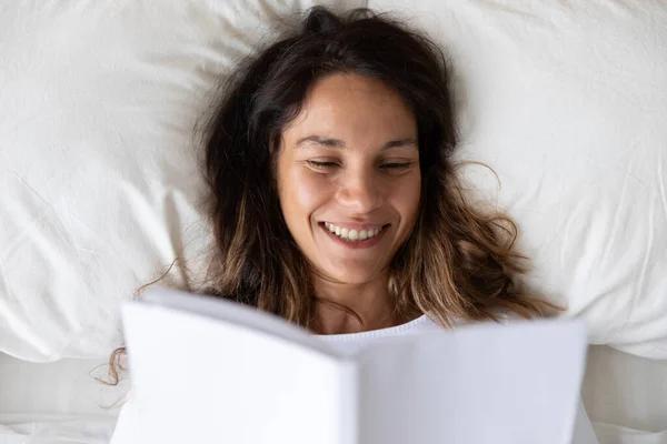 Улыбающаяся женщина читает книгу, отдыхая в постели — стоковое фото