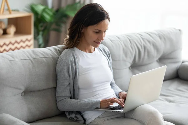 Mujer joven positiva usando el ordenador portátil, sentado en un sofá acogedor — Foto de Stock