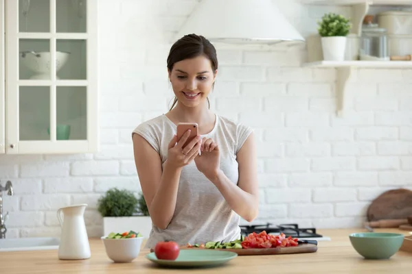 Lachende jonge aantrekkelijke vrouw koken naar internet recept op smartphone. — Stockfoto