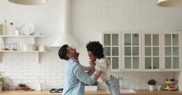 Happy caucasian dad lifting small african daughter playing in kitchen — Stock Video