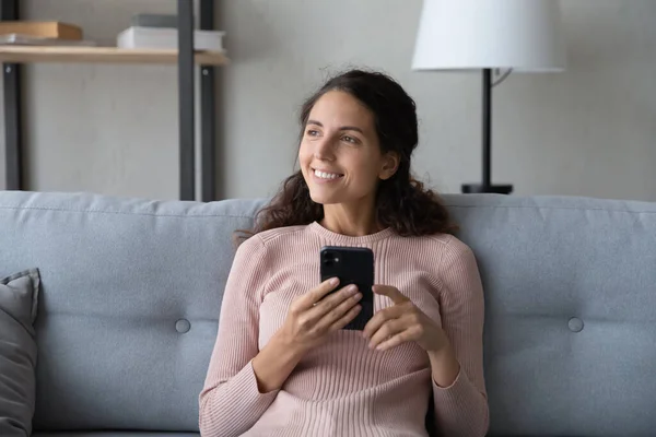 Distracted young smiling woman resting on sofa, holding smartphone. — Stock Photo, Image