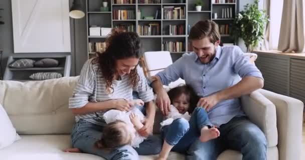 Счастливые молодые родители веселятся, щекоча маленьких детей на диване — стоковое видео