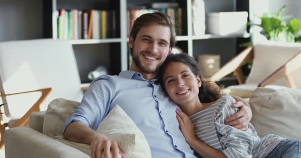 Sorrindo jovem casal abraçando no sofá confortável olhando para a câmera — Vídeo de Stock