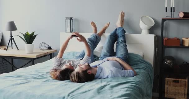 Despreocupado jovem casal relaxante deitado na cama usando smartphone moderno — Vídeo de Stock