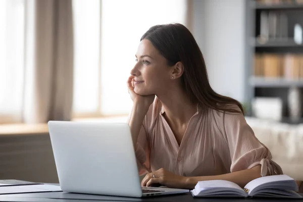 Jovem sonhadora ponderando ideias, sentada na mesa com laptop — Fotografia de Stock