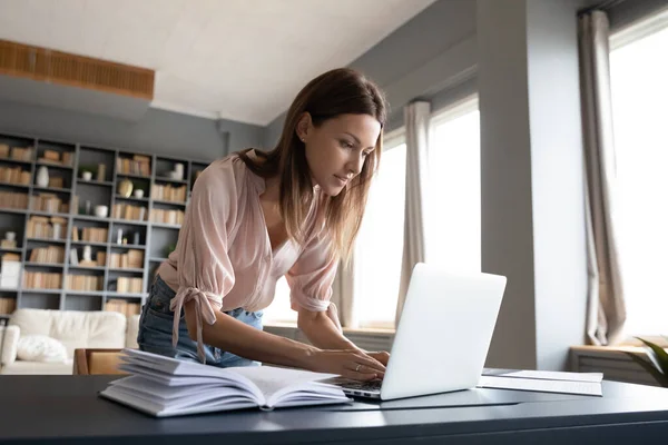 Leende kvinna står nära skrivbordet, med bärbar dator hemma — Stockfoto