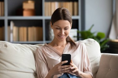 Gülümseyen genç bir kadın telefon kullanıyor, evdeki kanepede oturuyor.