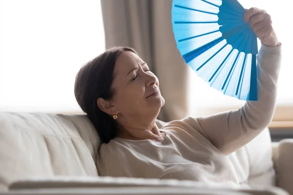 Cansado mulher de meia idade acenando ventilador azul, sofrendo de calor — Fotografia de Stock