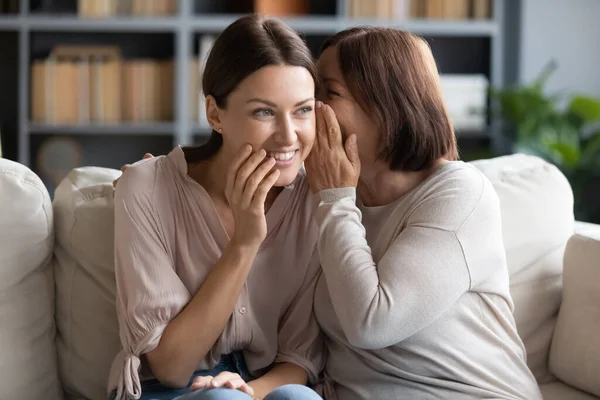 Middle aged mother whispering in smiling adult daughter ear, gossiping