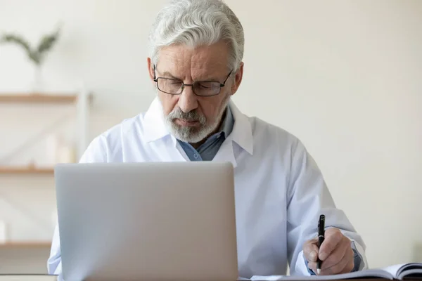 Tıp bürosunda dizüstü bilgisayar kullanarak notlar yazan yaşlı ciddi bir doktor. — Stok fotoğraf