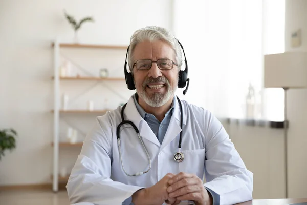 Glimlachende senior arts draagt headset kijkend naar camera, telegeneeskunde concept — Stockfoto
