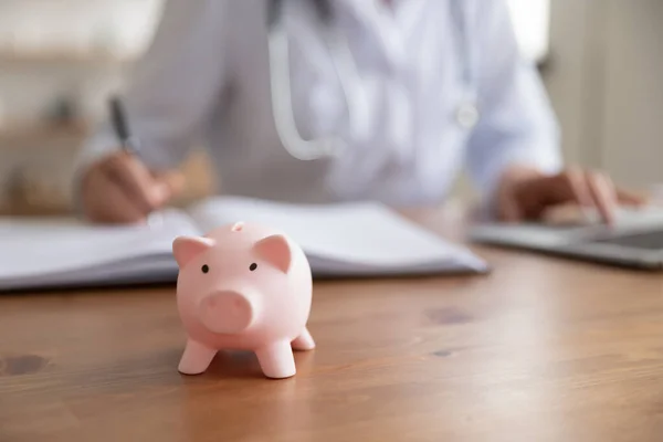 Ärztin arbeitet am Schreibtisch mit Sparschwein, Nahaufnahme — Stockfoto