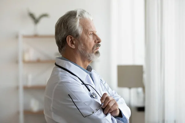 Задумчивый серьезный старший врач смотрит в окно, затерянное в мыслях — стоковое фото
