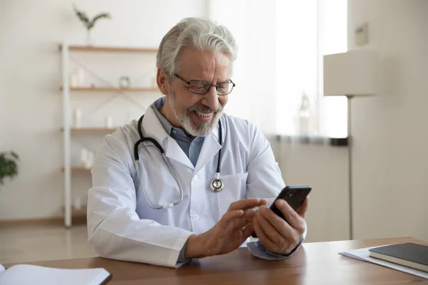 Feliz viejo médico sosteniendo el uso de teléfono inteligente en el consultorio médico —  Fotos de Stock