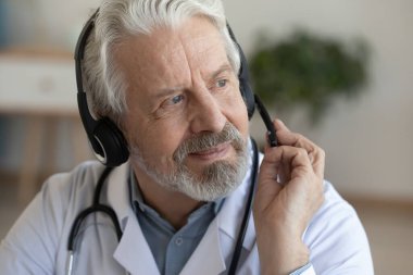 Mikrofonlu kulaklık takmış gülümseyen yaşlı doktor başka tarafa bakıyor.