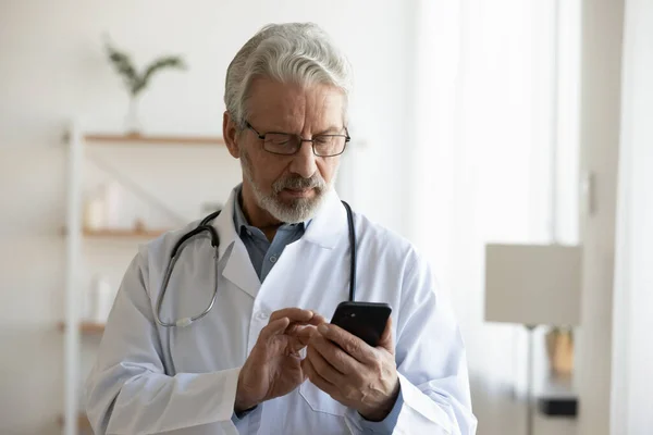 Médico varón mayor sosteniendo el teléfono inteligente usando aplicaciones médicas —  Fotos de Stock