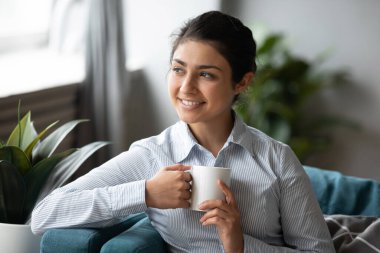 Smiling young indian woman relaxing on comfortable sofa, drinking coffee. clipart