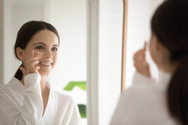 Jovem mulher olhar no espelho aplicar creme facial — Fotografia de Stock