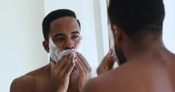 Homem africano aplicando espuma de barbear no rosto olhando no espelho — Vídeo de Stock