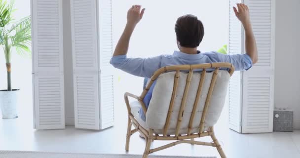 Hombre joven y tranquilo relajándose en una cómoda silla de madera disfrutando del salón — Vídeos de Stock