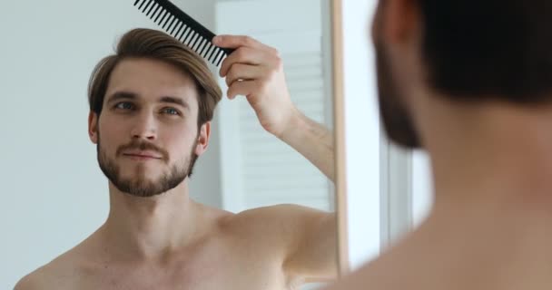 Bonito jovem escovar cabelo segurando pente olhando no espelho — Vídeo de Stock