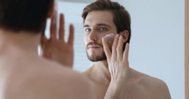 Yakışıklı genç bir adam aynaya bakarak yüz kremi sürüyor.