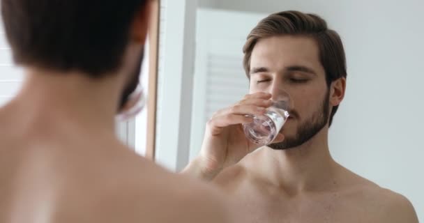 Schöner junger gesunder Mann, der Wasser trinkt und in den Spiegel schaut — Stockvideo