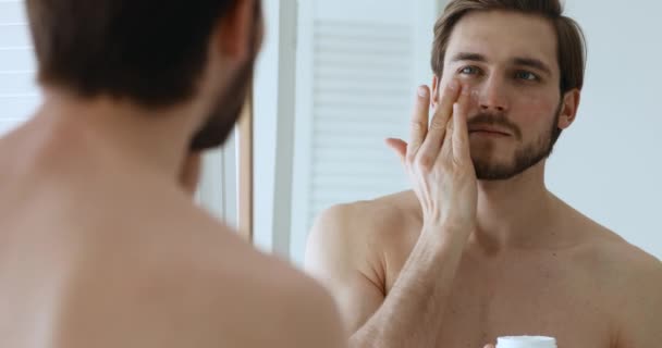 Attractive young guy applying cream on face looking in mirror — Stock Video