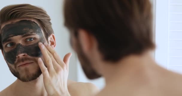 Sonriente joven metrosexual hombre aplicando máscara facial mirando en el espejo — Vídeos de Stock