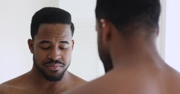 Улыбающийся африканский мужчина очищает кожу лица, глядя в зеркало ванной — стоковое видео