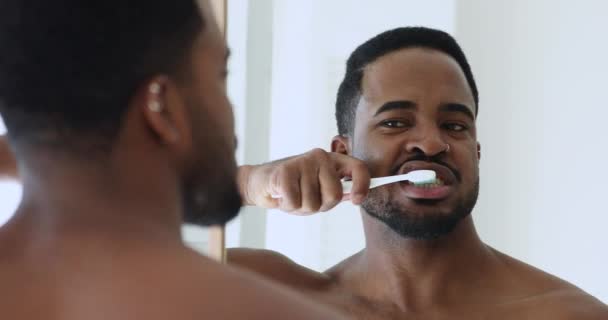 Jovem homem africano segurando escova de dentes escovando os dentes olhando no espelho — Vídeo de Stock