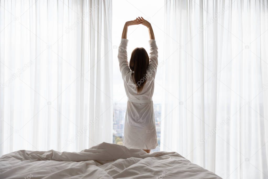 Rear view woman standing near window raised hands stretching body