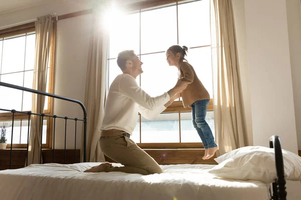 Happy father playing with little daughter, jumping on bed