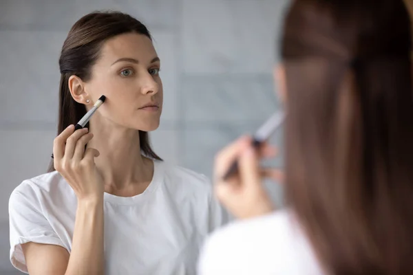 Woman reflected in mirror holding brush applying foundation concealer cream