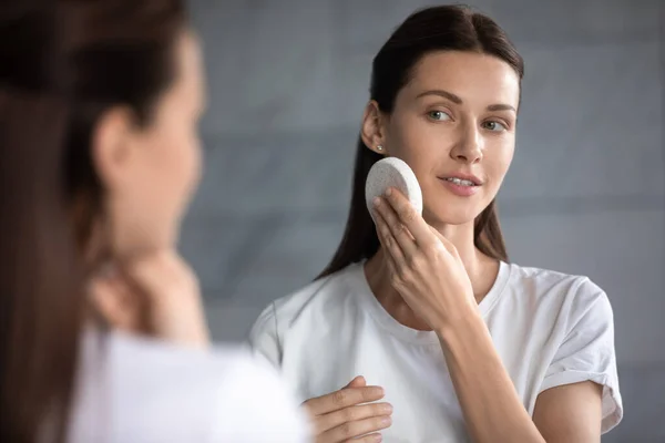 Mulher refletindo no espelho segurando rosto esponja facial limpa — Fotografia de Stock