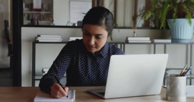 İşyerinde dizüstü bilgisayar kullanarak notlar alan odaklanmış genç Hintli işçi.